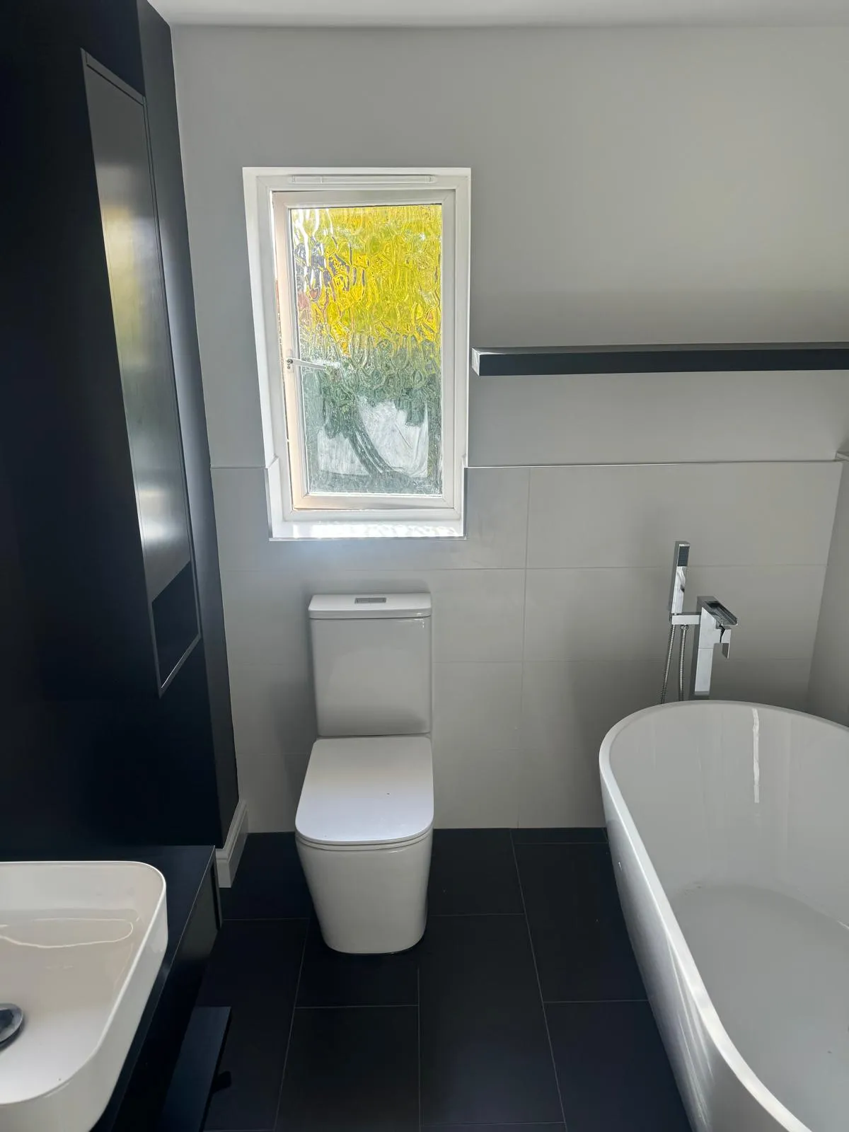 A white bathroom with a freestanding bath, a floor standing tap and a dark tiled floor