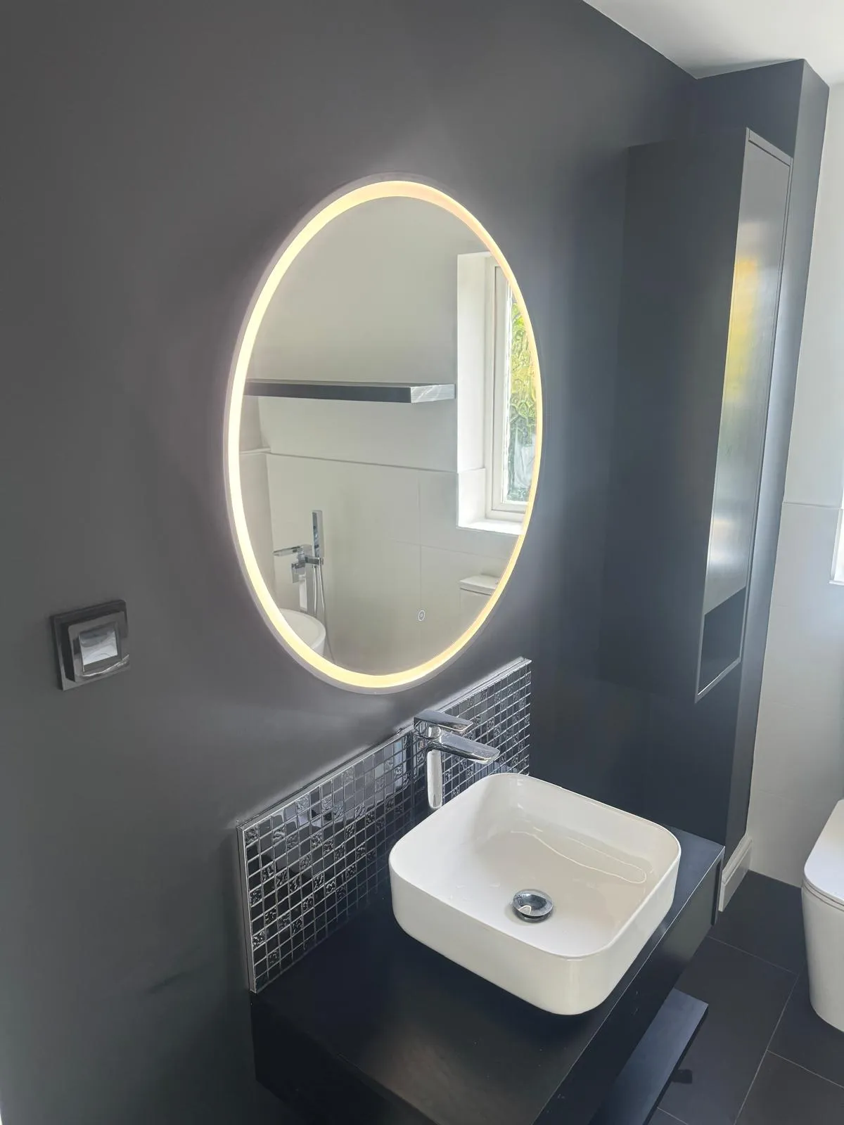 A square basin under a round lit mirror on a dark grey wall